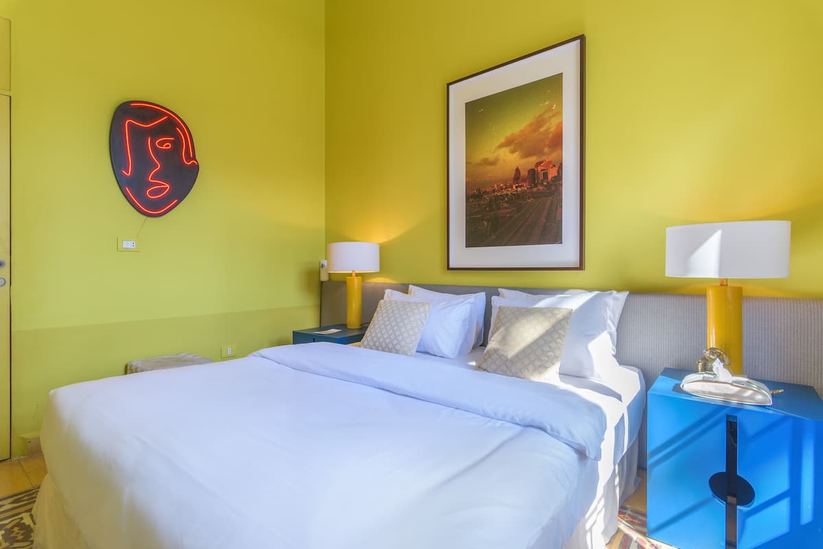 A hotel room with a bed, two bedside tables with lamps, a neon art piece, and a framed picture on the wall.