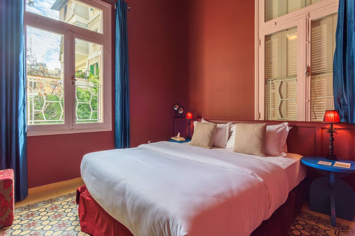 A hotel room with a large bed, a bedside table with a lamp, and a window with shutters.