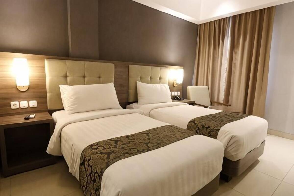 A hotel room with two beds, a nightstand, a lamp, and a view of the outside.