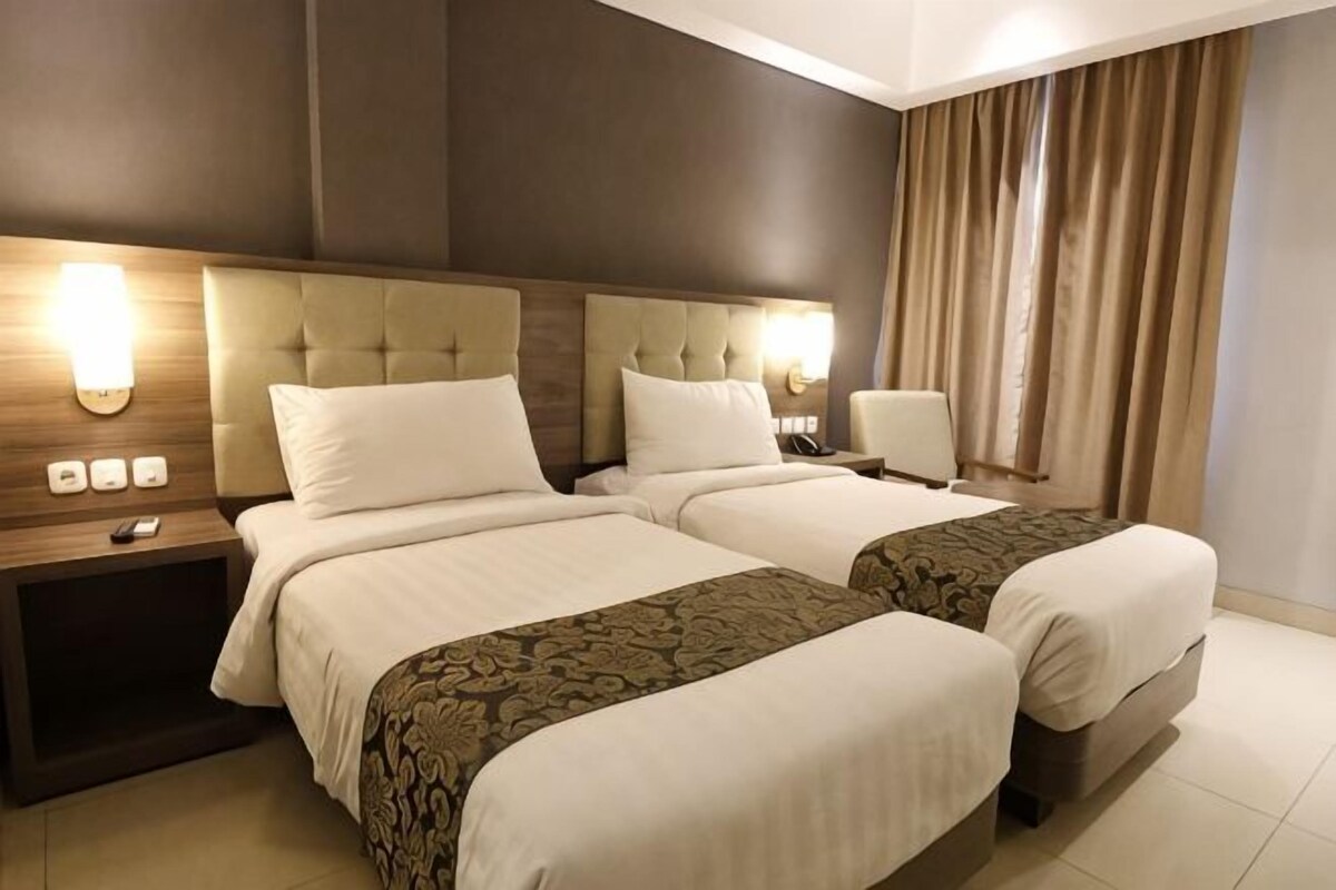 A hotel room with two beds, a nightstand, a lamp, and a view of the outside.