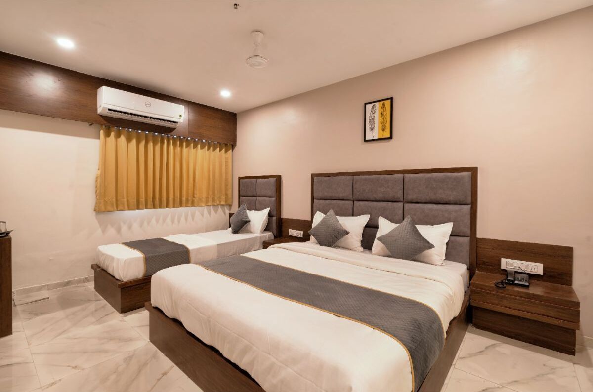A hotel room with two beds, a large window with curtains, an air conditioning unit, a bedside table with a phone, and a wall-mounted artwork.