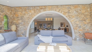 A modern living room with a stone wall, a sofa, a coffee table, and a dining area in the background.