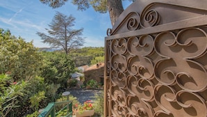 A wrought iron gate with intricate scrollwork, leading to a courtyard with a stone house, lush greenery, and a clear blue sky.