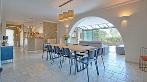 A spacious dining area with a long wooden table and multiple chairs, a kitchen area with a bar, and a large arched window providing natural light.