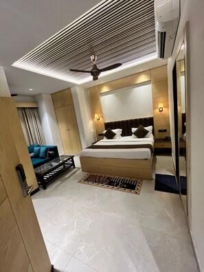 A modern hotel room with a large bed, a sofa, a ceiling fan, and a large mirror. - Hotel sar kamla dham (Mathura)