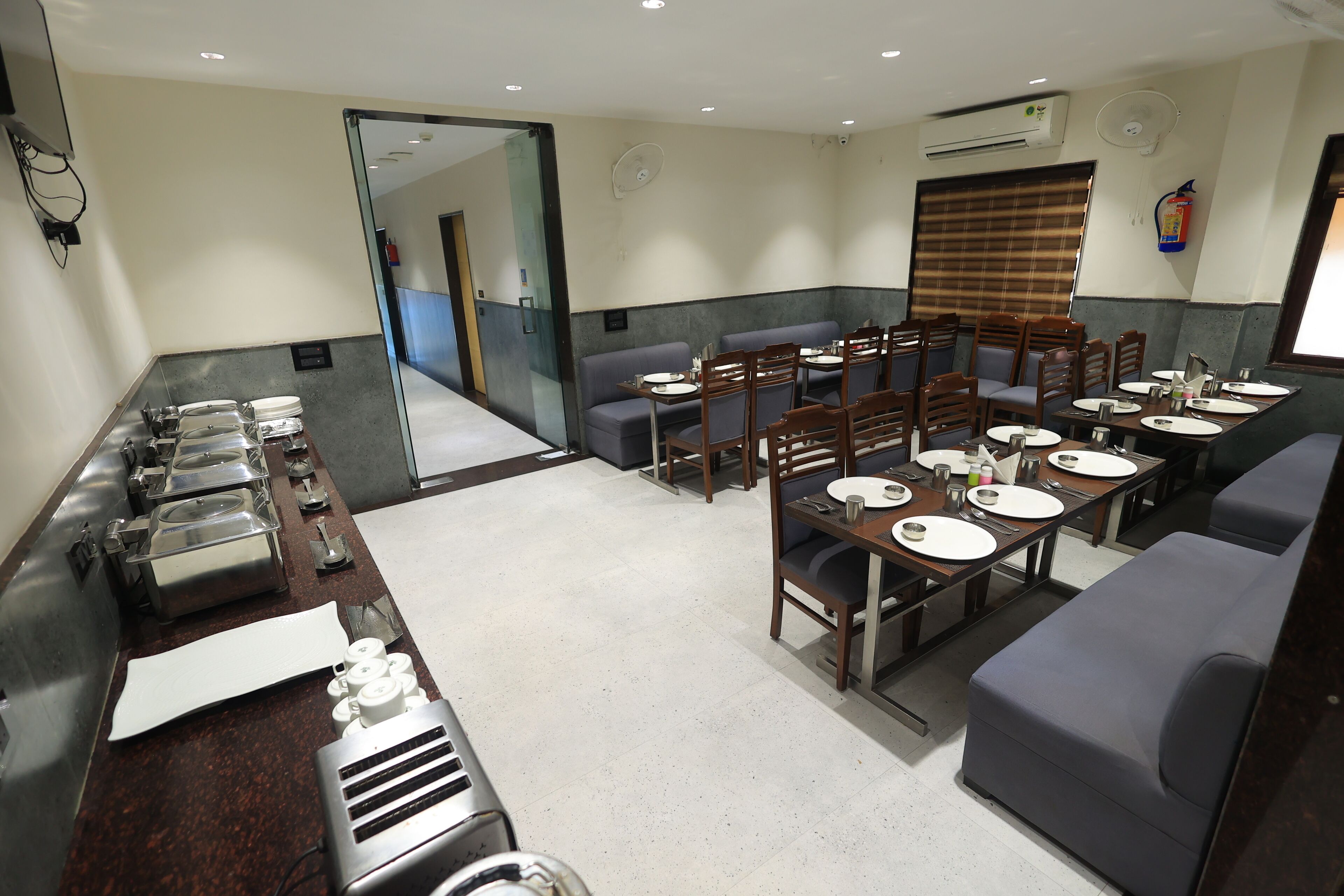 A hotel dining area with multiple tables and chairs, a buffet counter with food items, and a fire extinguisher on the wall.