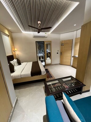 A modern hotel room with a bed, a glass coffee table, a sofa, a ceiling fan, and a wardrobe. - Hotel sar kamla dham (Mathura)
