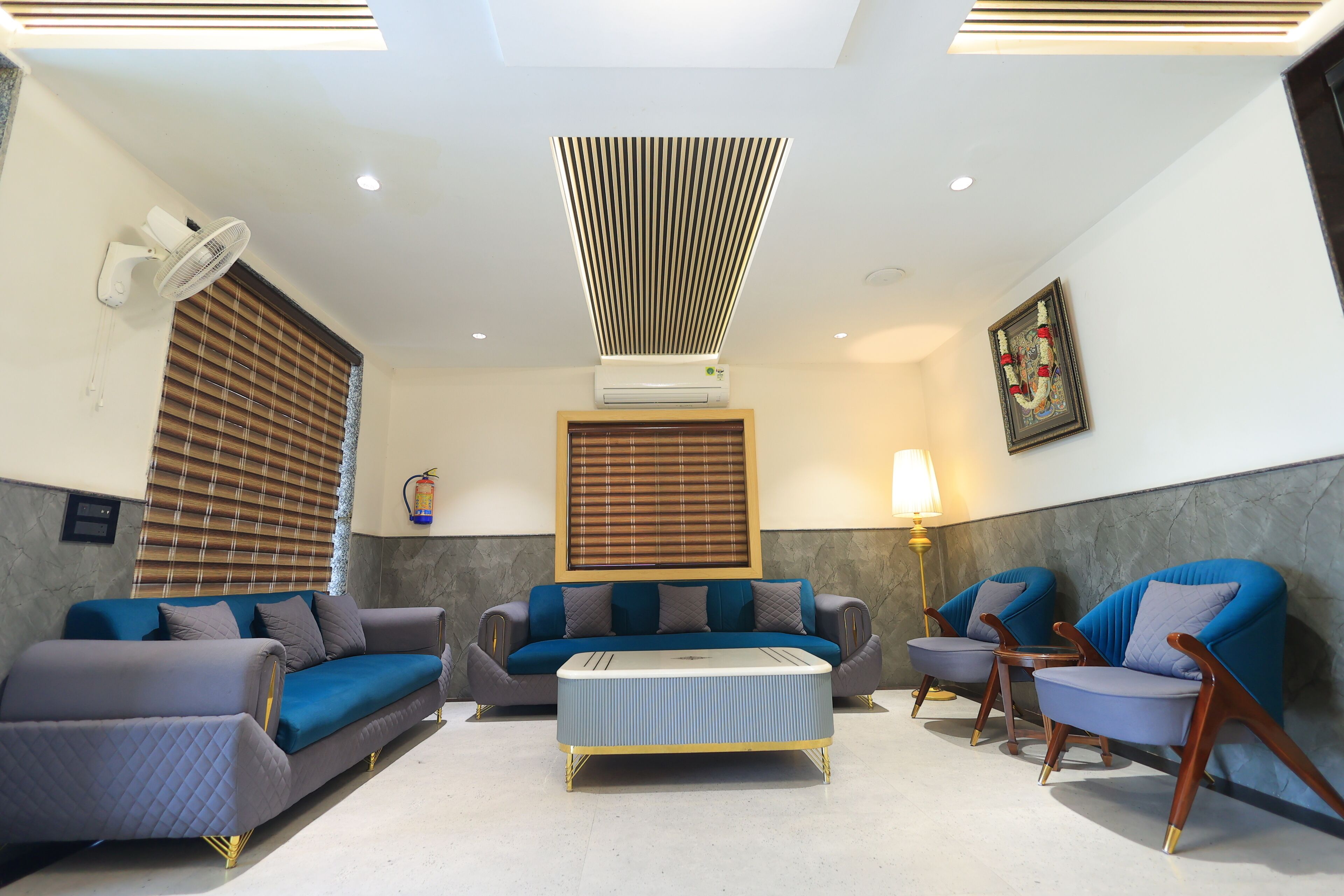 A modern living room with a sofa, two armchairs, a coffee table, a standing fan, an air conditioning unit, a wall-mounted lamp, and a framed picture.