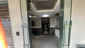 Um saguão de hotel com entrada por porta de vidro, um sofá, uma mesa de centro e um quadro na parede.