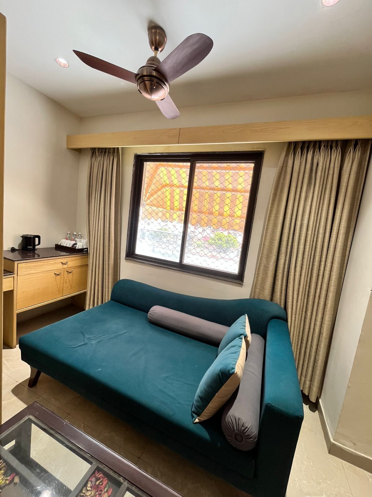 A room with a teal sofa, a ceiling fan, a window with curtains, a small table with a coffee maker, and a wooden desk.