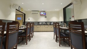 A dining area with wooden chairs and tables, an air conditioning unit, a clock, and a television screen. - Hotel sar kamla dham (Mathura)