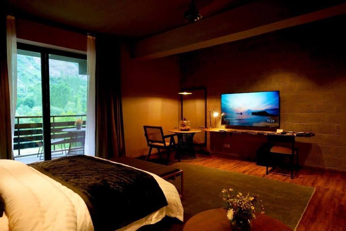 Quarto de hotel com uma cama grande, uma televisão em um suporte, uma mesinha com um vaso, e uma varanda com vista para o verde.