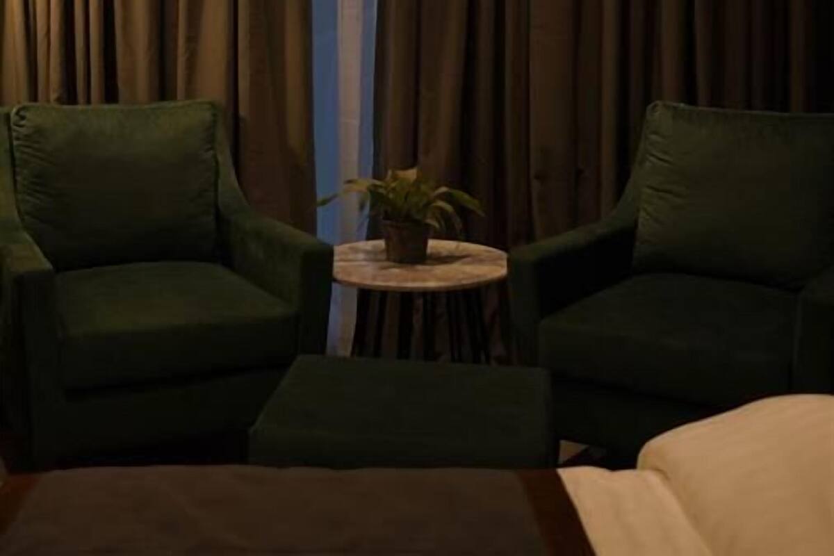 Quarto com duas poltronas verdes, uma mesinha redonda com uma planta e uma janela grande com cortinas.