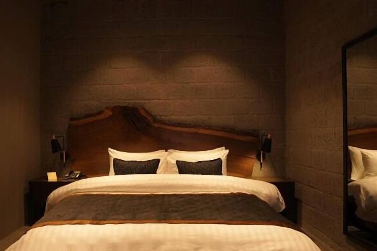 Quarto de hotel com cabeceira de madeira, cama com lençóis brancos, mesa de cabeceira com abajur e quadro pendurado na parede.