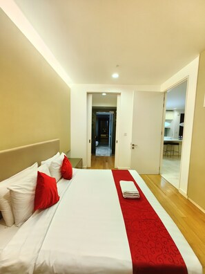 A hotel room with a large bed, white bedding, red pillows, and a red patterned runner. There is a wooden floor and a door leading to a hallway and a kitchen area. - Amazing Pool By Platinum Suites (Kuala Lumpur)