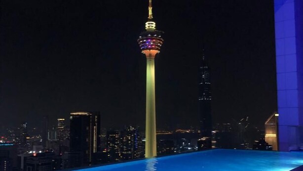 Ein Blick auf ein Schwimmbecken bei Nacht, im Hintergrund eine Stadtlandschaft und ein Turm.