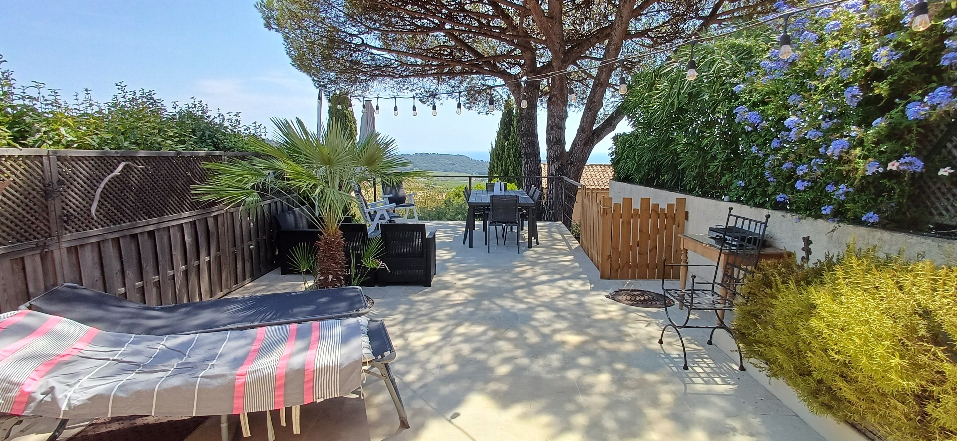An outdoor patio area with a dining table and chairs, surrounded by a wooden fence and various plants.