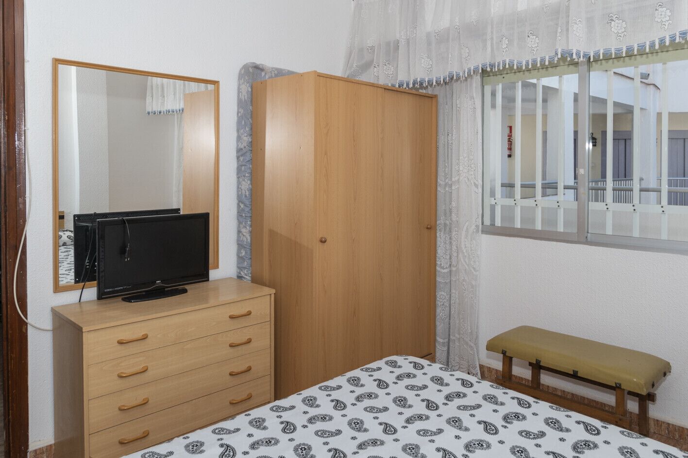 A bedroom with a wooden wardrobe, a bed with a patterned bedspread, a bench, a television on a dresser, and a window with a view of the outside.