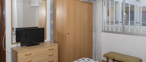 A bedroom with a wooden wardrobe, a bed with a patterned bedspread, a bench, a television on a dresser, and a window with a view of the outside.