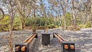 An outdoor seating area with benches and a central table, surrounded by trees and gravel.