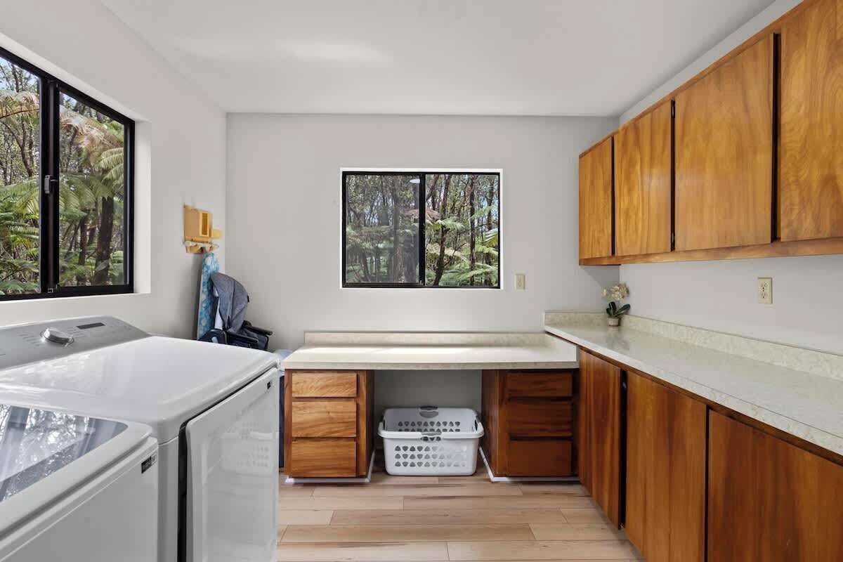 A compact kitchen with wooden cabinets, a built-in washing machine, and a window with a forest view.