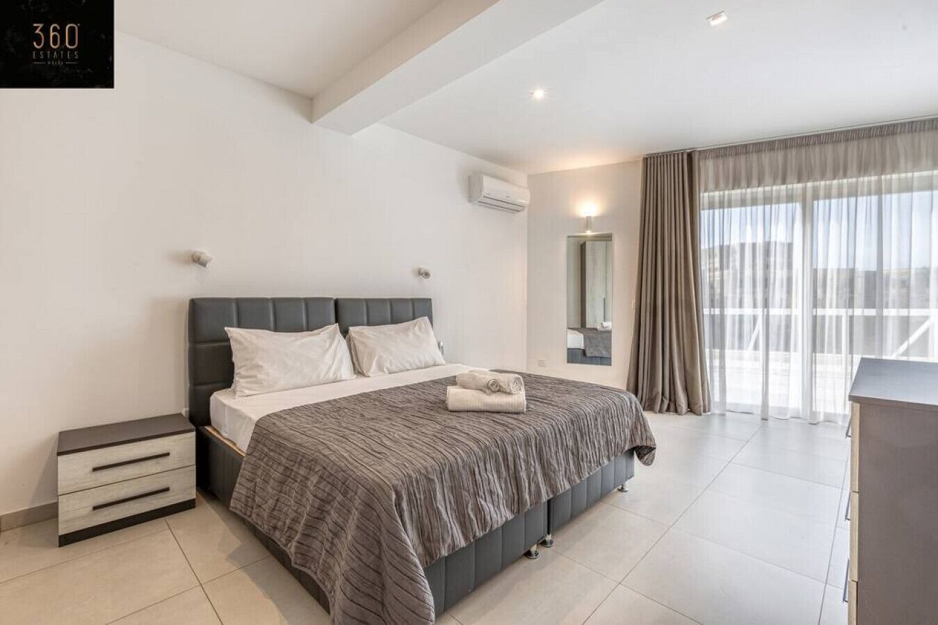 Un dormitorio moderno con una cama grande, una mesita de noche, un aparador y un amplio ventanal con cortinas.