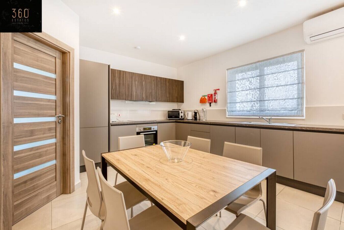 A modern kitchen with a wooden table, white chairs, and a window with blinds.