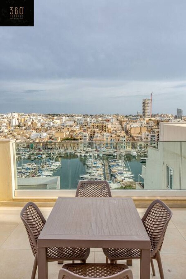 Un balcón con mesa y sillas da a un puerto deportivo con barcos y vistas de la ciudad.