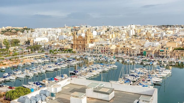 Un puerto deportivo lleno de barcos, un paisaje urbano con edificios históricos y un cielo despejado.