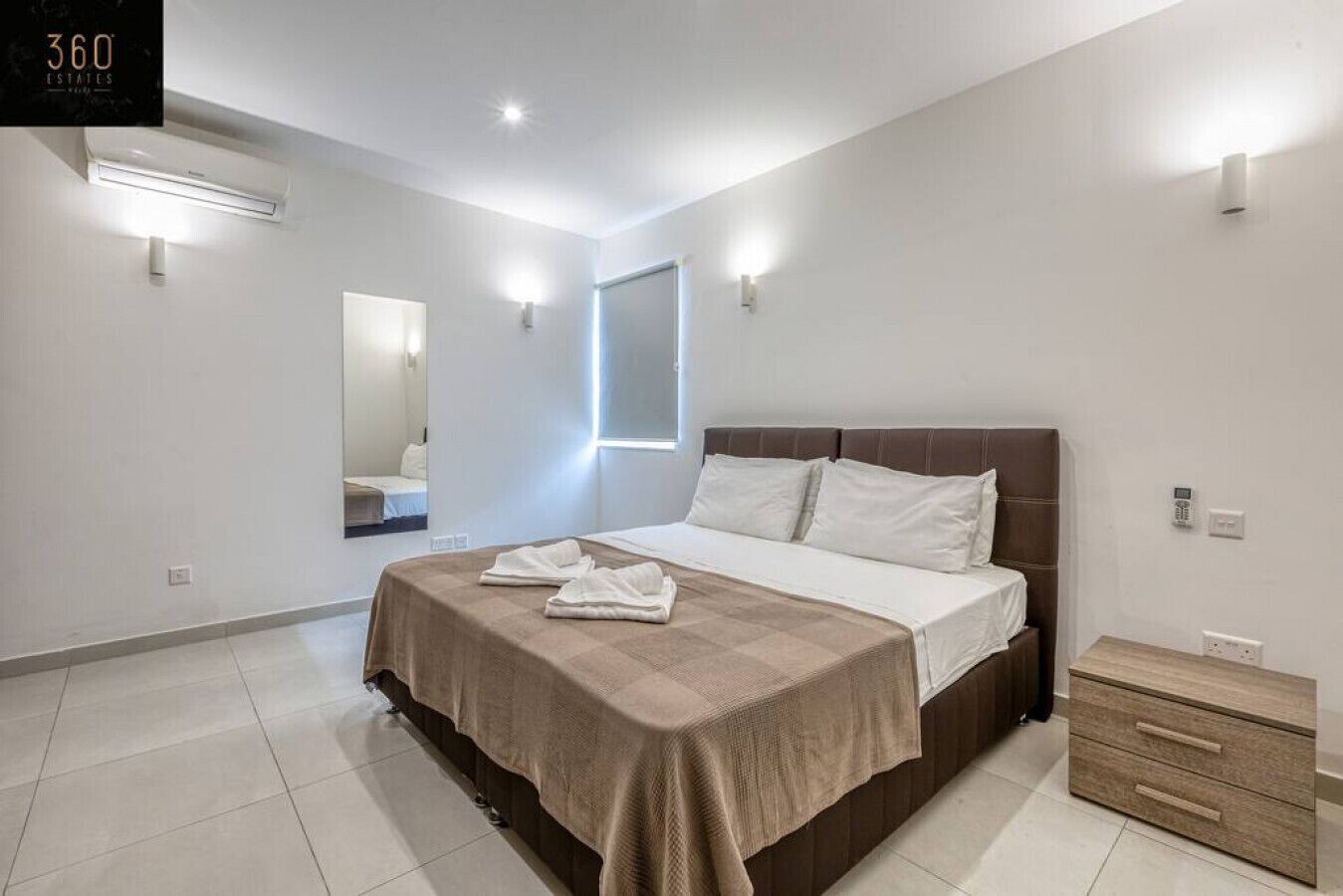 Un dormitorio moderno con una cama grande, mesitas de noche, un espejo e iluminación en la pared.