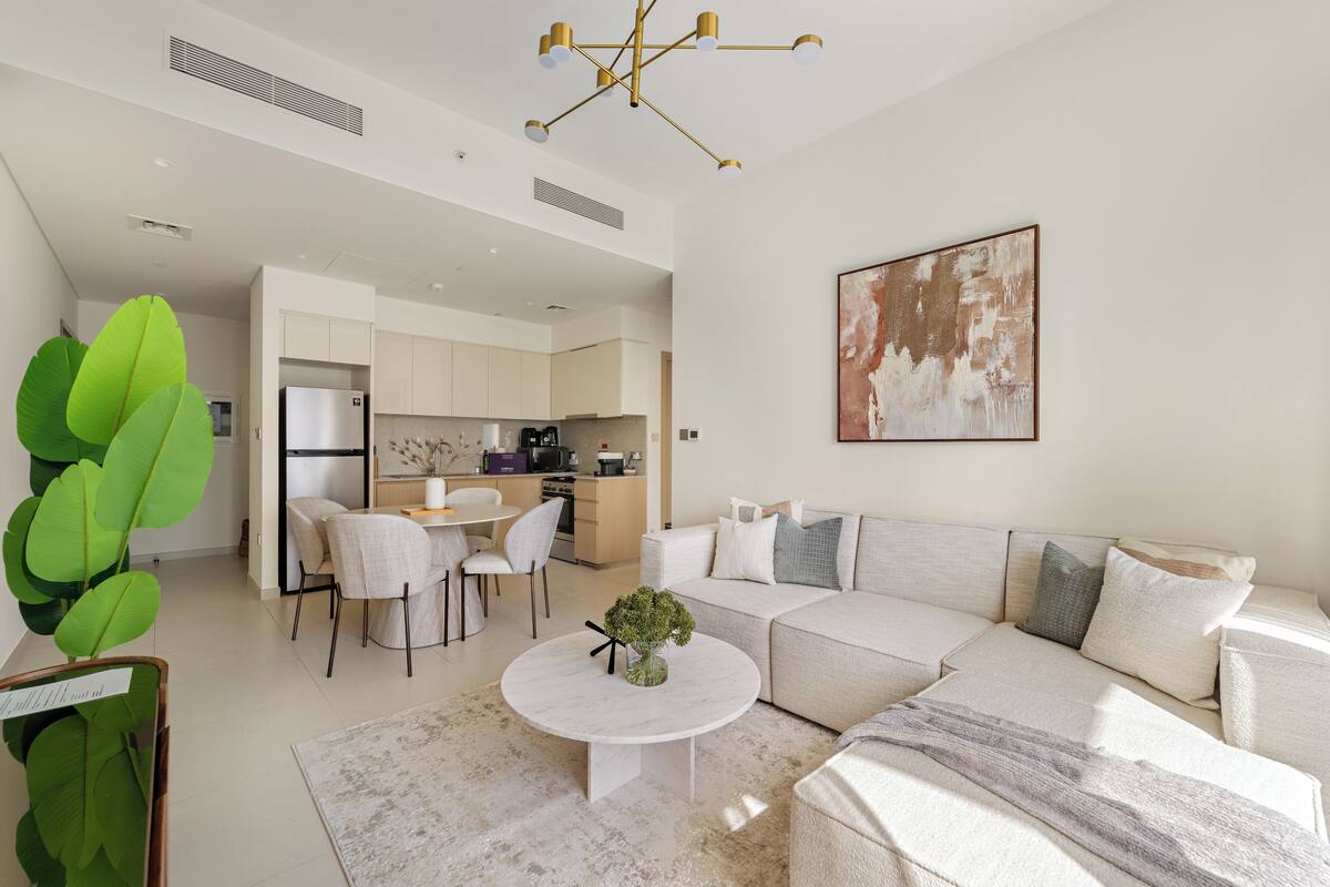 A modern living room with a sectional sofa, a dining area with chairs, a kitchen in the background, and a large abstract painting on the wall.