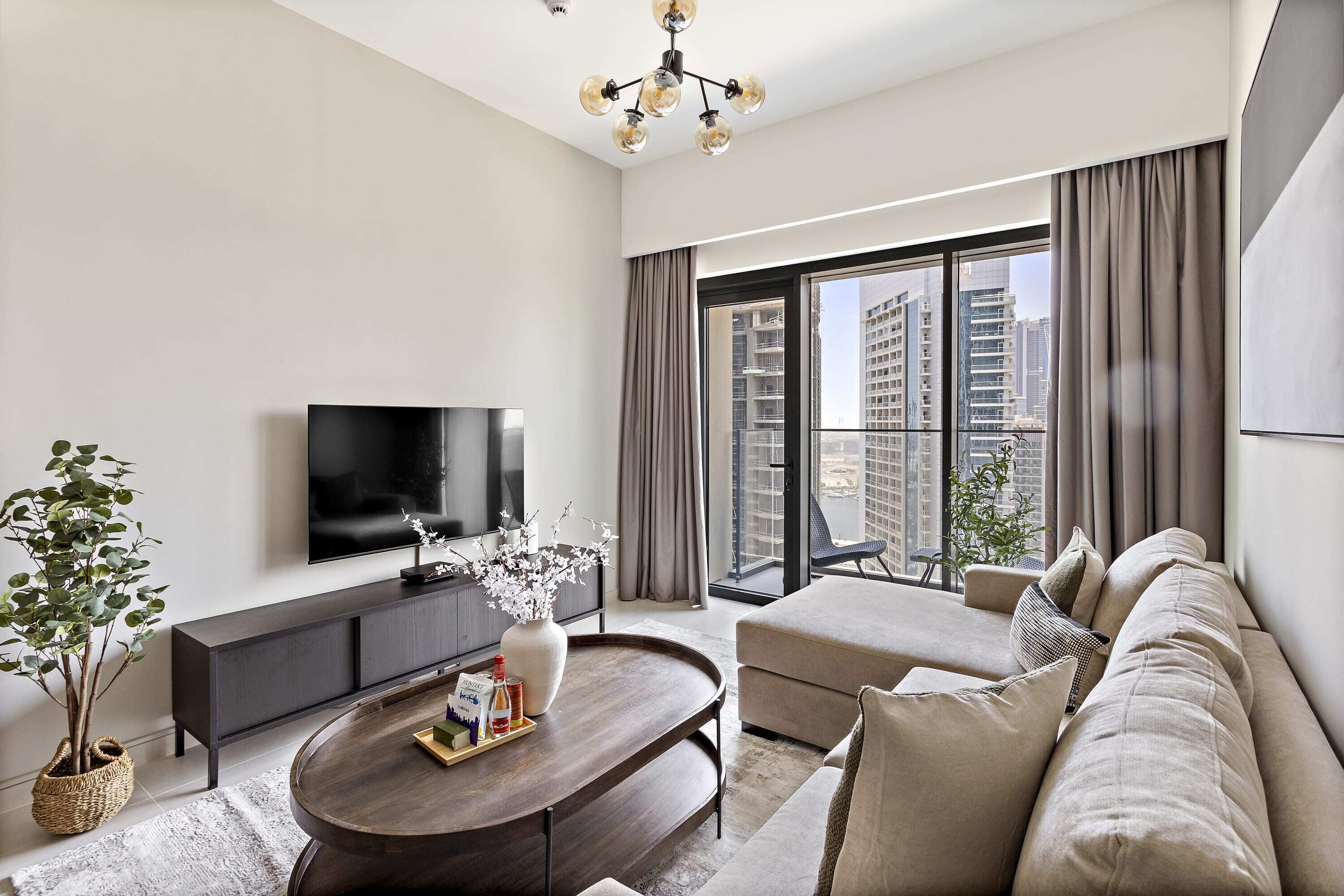 A modern living room with a flat-screen TV, a dark wood coffee table, a beige sofa, and a large window offering a city view.