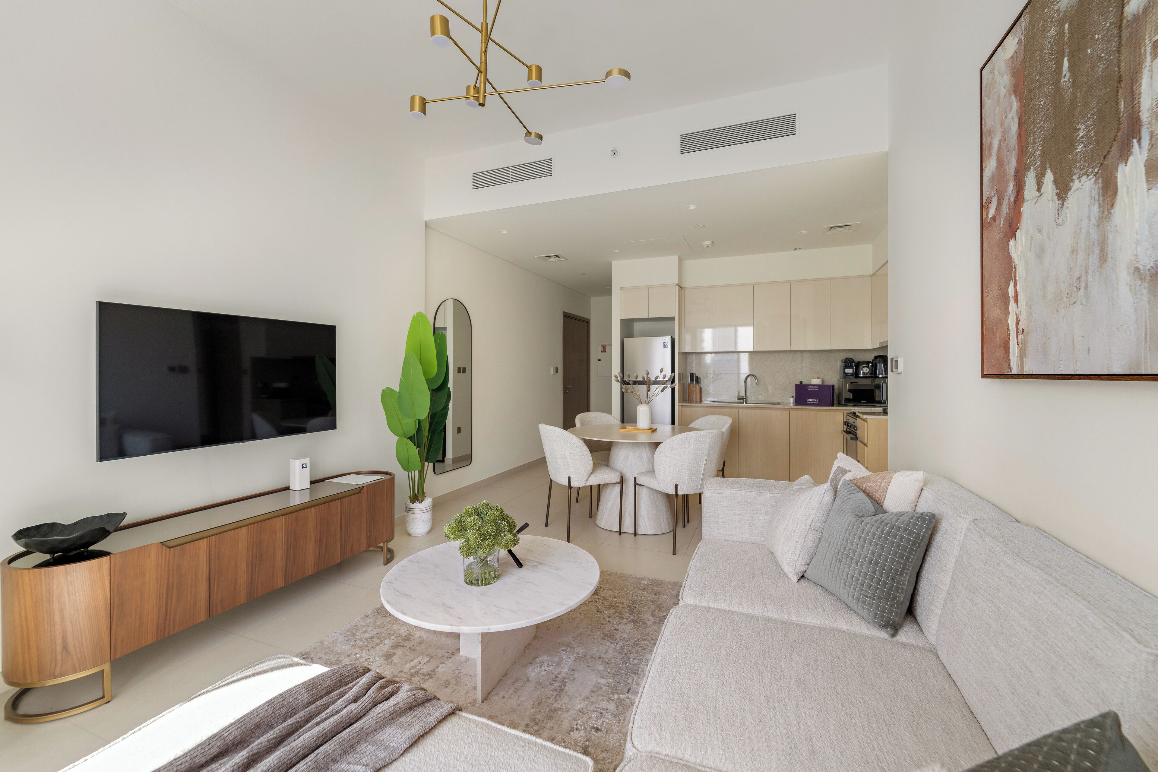 Ein modernes Wohnzimmer mit einem Flachbildfernseher, einer Holzkonsole, einem runden Couchtisch mit einer Pflanze, einem grauen Sofa und einem Essbereich mit weißem Tisch und Stühlen.