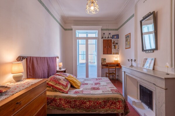 A bedroom with a bed, dresser, desk, fireplace, and a door leading to an outdoor balcony.