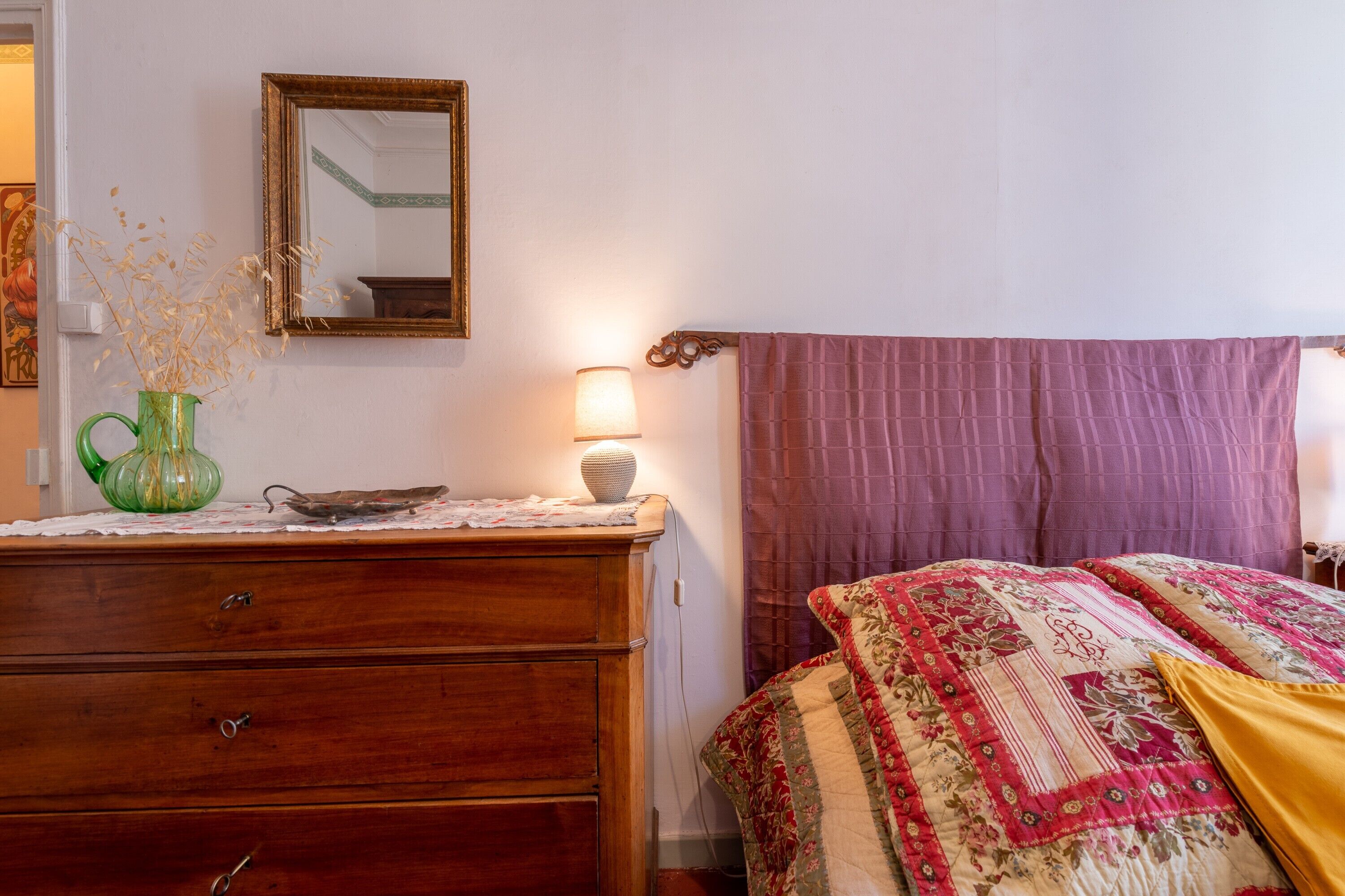 A bedroom with a wooden dresser, a mirror, a lamp, a bed with a floral bedspread, and a purple curtain.