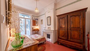 Une pièce avec une armoire en bois, une cheminée, un lit recouvert d’une couvre-lit à motifs floraux, un vase vert et une fenêtre donnant sur un beau paysage.