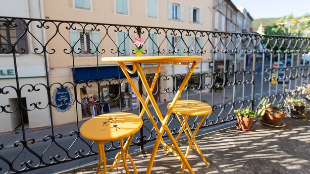 Une table et des chaises en métal jaune sur un balcon agrémenté d’une balustrade en fer forgé, de plantes en pot et, en arrière-plan, d’un immeuble aux murs et aux volets de couleur claire.