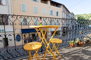 A yellow metal table and chairs on a balcony with a wrought iron railing, potted plants, and a building with light-colored walls and shutters in the background.