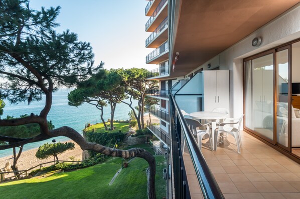 Un balcón con vista al mar, un área de comedor con sillas, y un edificio con varios pisos y balcones.