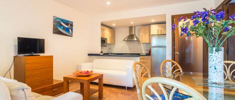 A living room with a sofa, TV, wooden cabinet, dining table, chairs, and a kitchen in the background.
