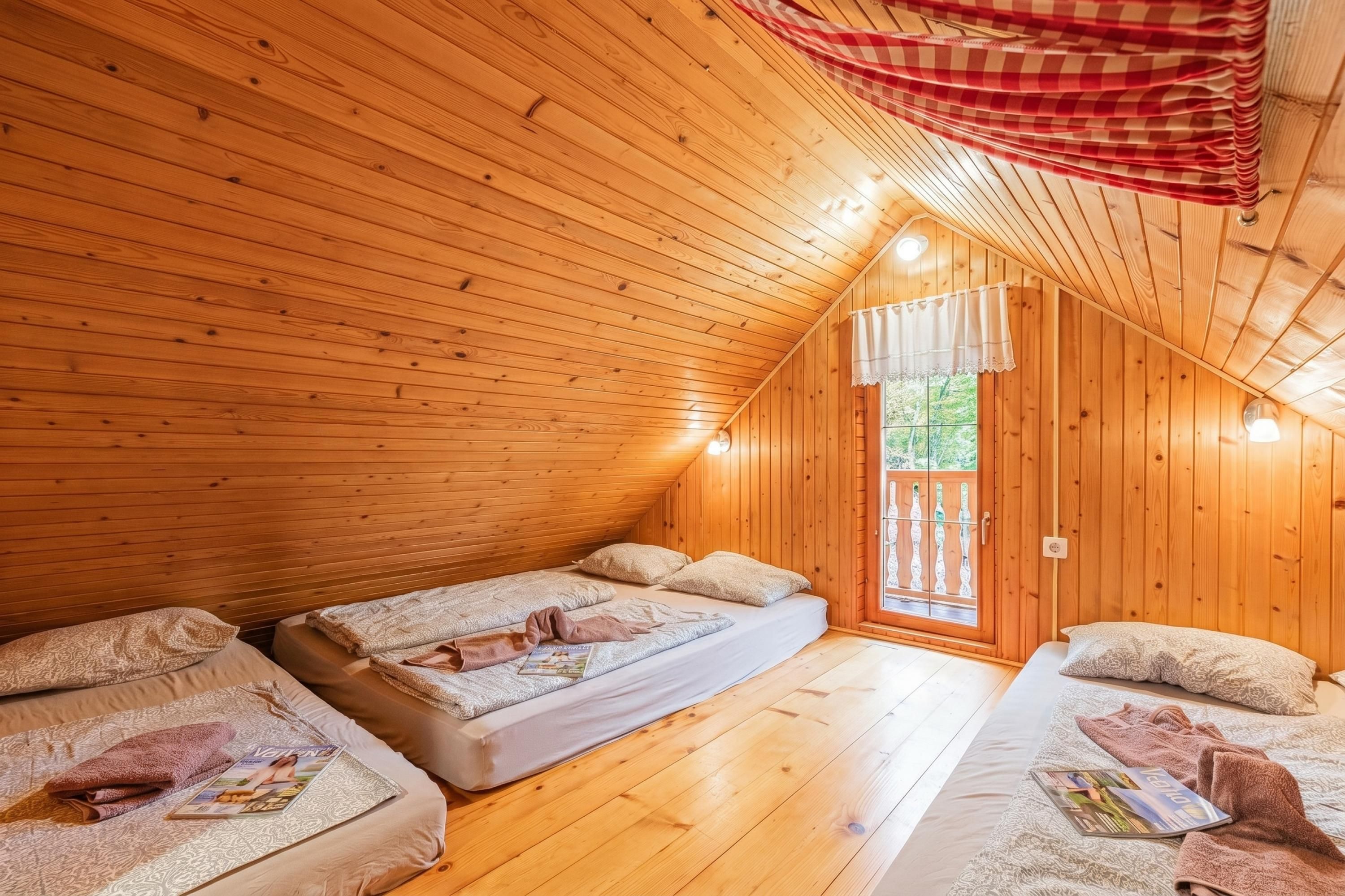 Una cabaña acogedora con paredes de madera y techo inclinado, que cuenta con dos camas con almohadas y mantas, un ventanal pequeño y una cortina roja a cuadros.