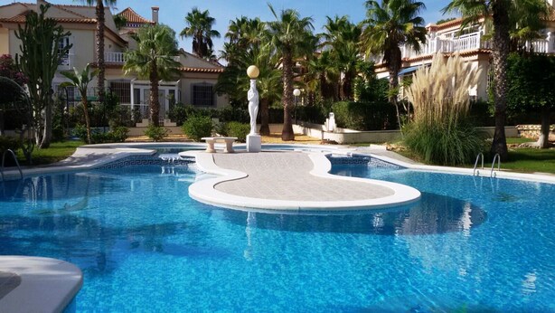 A swimming pool with a central island, surrounded by palm trees and a building with a tiled roof. - Confort Apartamento con Excelentes Vistas al mar (VC)