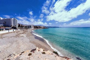 A beach with a clear blue sky, a sandy shore, and a building with multiple floors on the left.