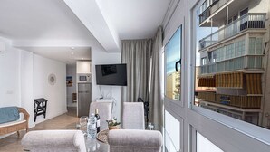Ein modernes Wohnzimmer mit einem Flachbildfernseher, grauen Sofas, einem Glastisch und Blick auf ein Gebäude mit Balkonen.