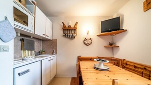 Une cuisine avec des meubles blancs, un four à micro-ondes, une table à manger en bois avec des bancs, un téléviseur fixé au mur et une étagère avec des objets décoratifs.