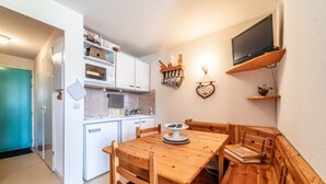 Une cuisine compacte équipée d’un four à micro-ondes, d’un four et de meubles blancs. On y trouve une table à manger en bois avec des bancs, une petite télévision posée sur une étagère et une décoration en forme de cœur accrochée au mur.