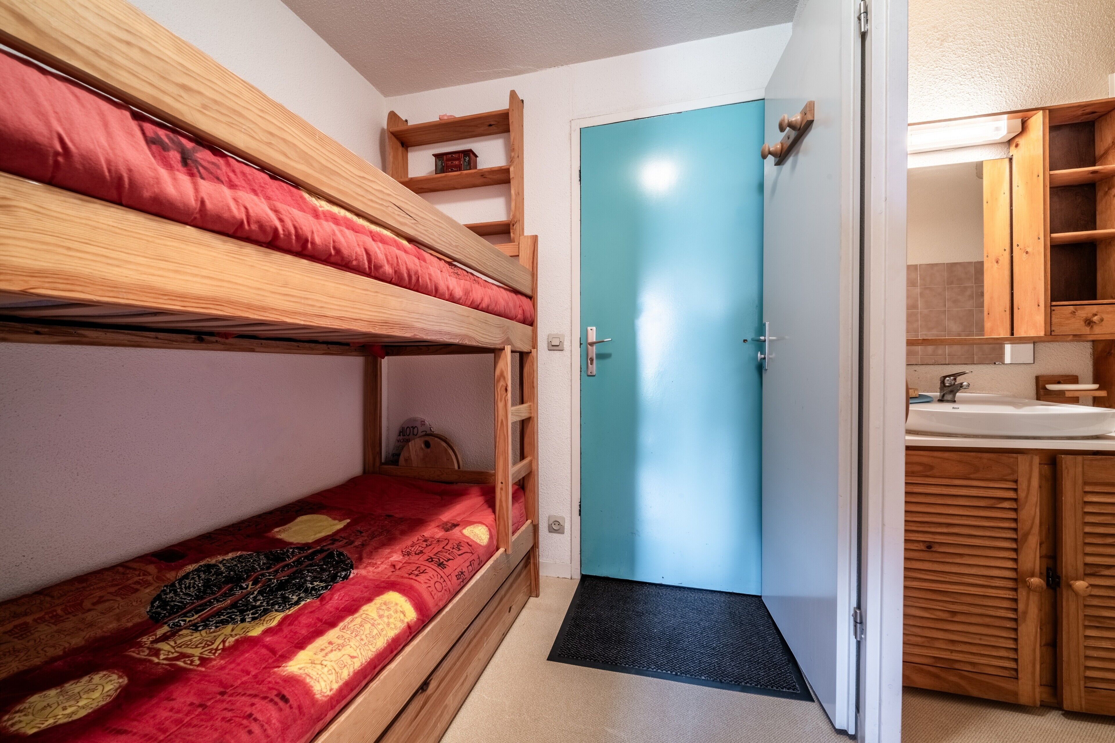 A bunk bed with a red blanket, a blue door, and a wooden cabinet with a mirror.