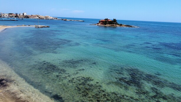 A coastal area with clear turquoise waters, a sandy beach, and a small island with a building.