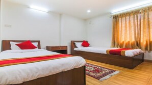 A hotel room with two single beds, a small bedside table, a nightstand, a red and white bedspread, a red and gold throw blanket, a patterned rug, and a window with curtains. - MeroStay 186 Hotel RR (Kathmandu)