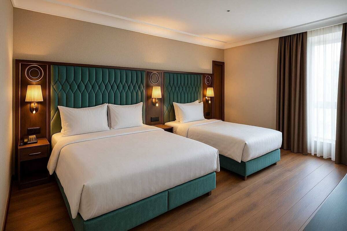 Une chambre d’hôtel avec deux lits, une tête de lit recouverte d’un tissu vert, des lampes de chevet et une grande fenêtre avec des rideaux.